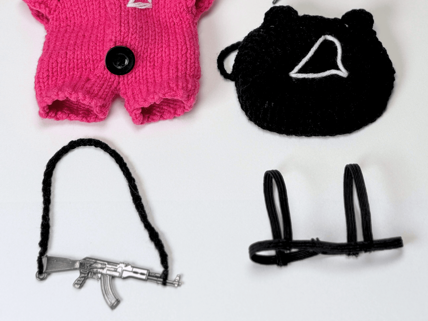 Detail shot of handcrafted Squid Game pink guard doll clothes set including knitted mask, gun prop with crochet strap, and harness