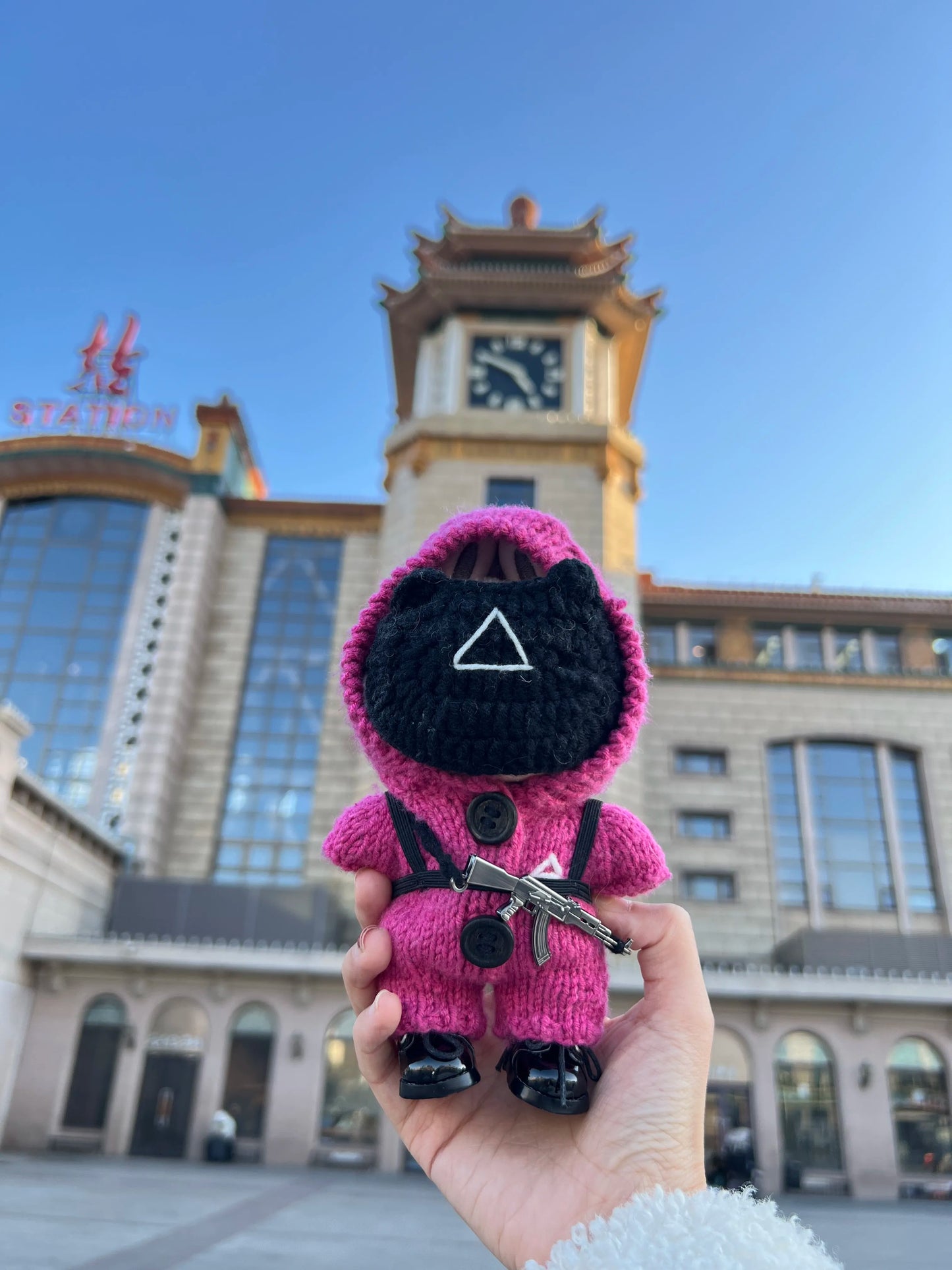 Pink guard Labubu toy with a black mask held in front of a clock tower and building.