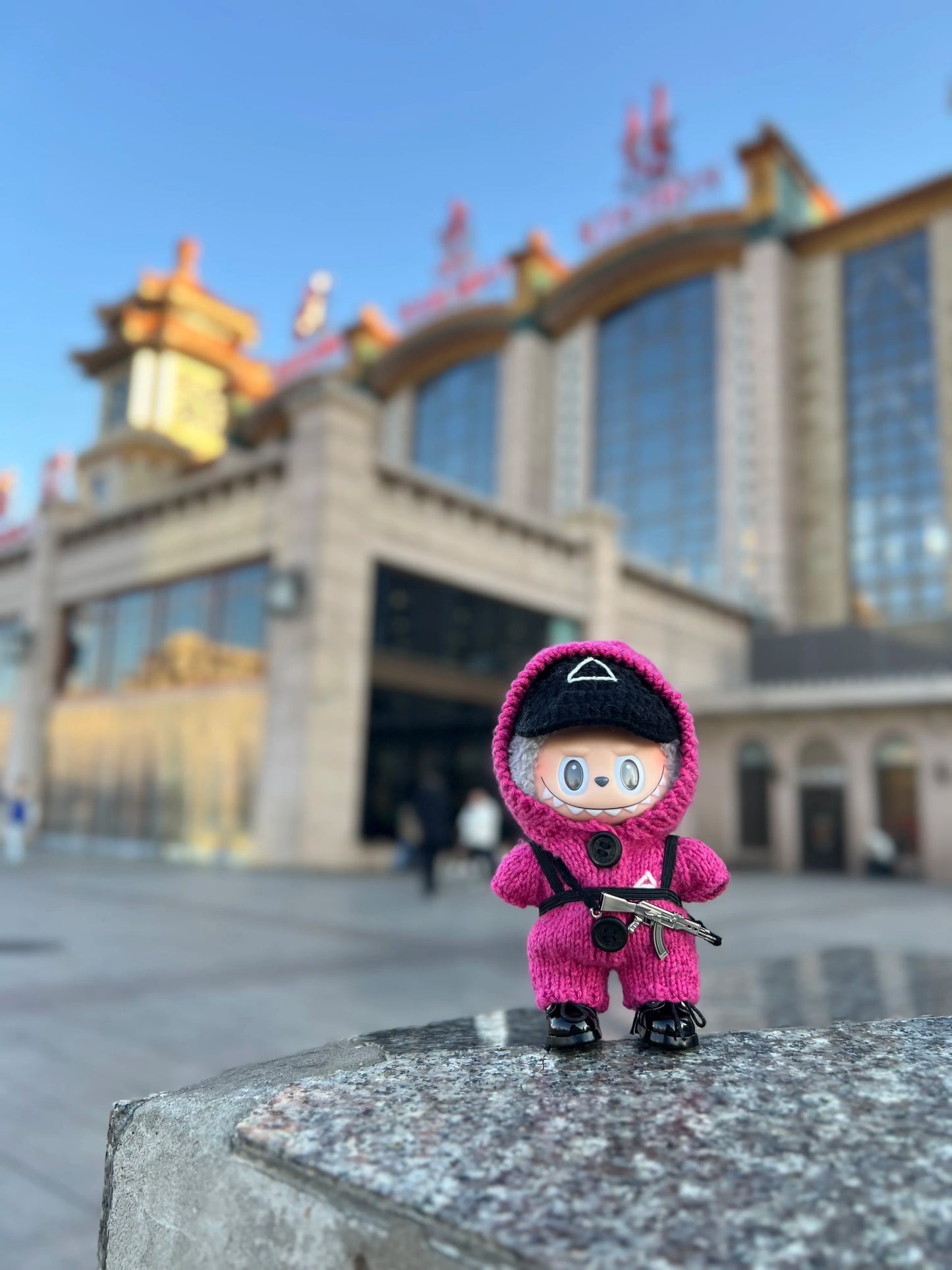 Pink guard Labubu toy with a black mask standing in front of a clock tower and building.