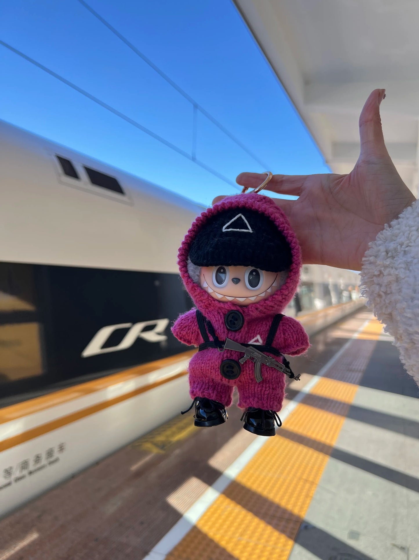 Plush Labubu wearing pink guard outfit held up against a train platform with a CR train in the background.