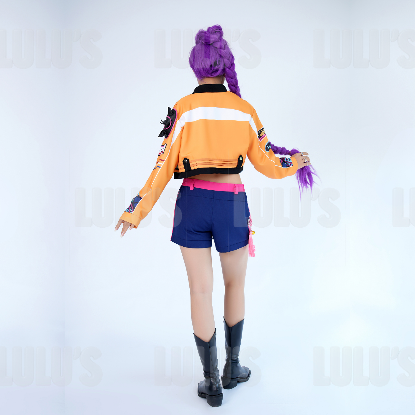 Model wearing K-Pop Demon Hunters Rumi cosplay costume with purple braided wig, orange cropped jacket, navy blue shorts, and black boots, shown from the back view.