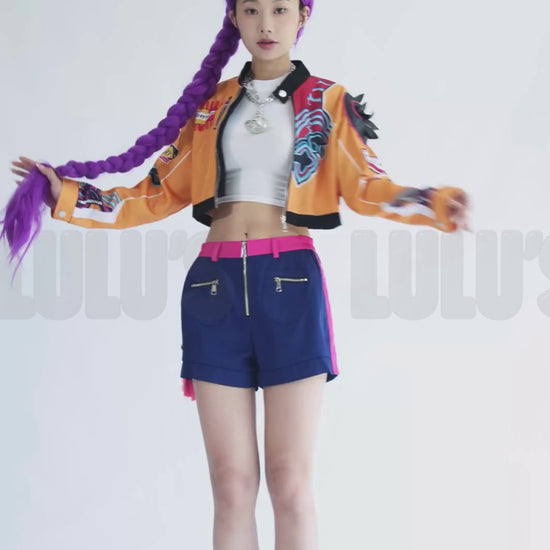 Video of female model posing in K-Pop Demon Hunters Rumi cosplay costume with purple wig, orange cropped jacket, navy shorts, pink belt, and black boots.