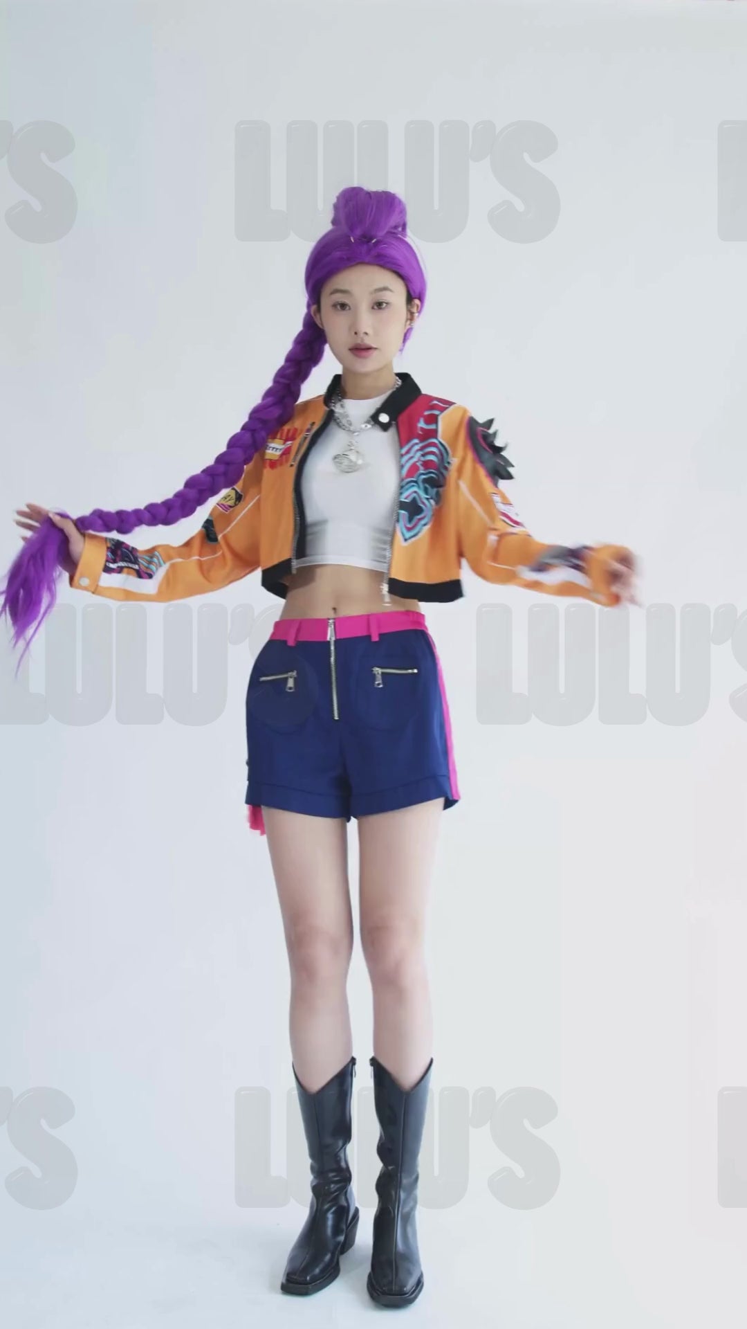 Video of female model posing in K-Pop Demon Hunters Rumi cosplay costume with purple wig, orange cropped jacket, navy shorts, pink belt, and black boots.