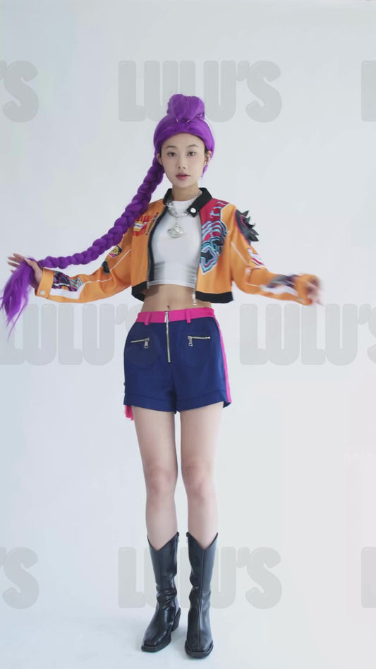 Video of female model posing in K-Pop Demon Hunters Rumi cosplay costume with purple wig, orange cropped jacket, navy shorts, pink belt, and black boots.